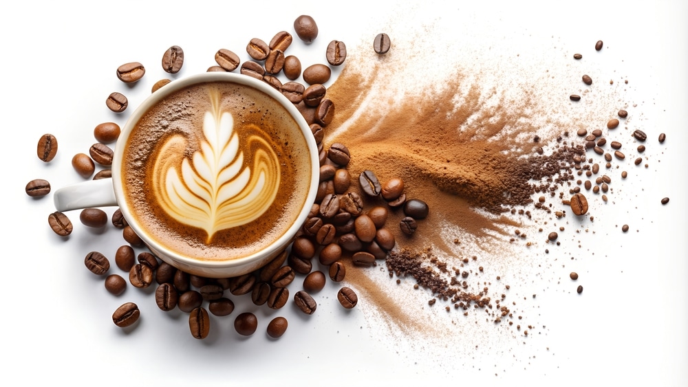 Bovenaanzicht van een kop cappuccino met latte-art in de vorm van een blad, op de tafel liggen losse koffiebonen en gemalen koffie