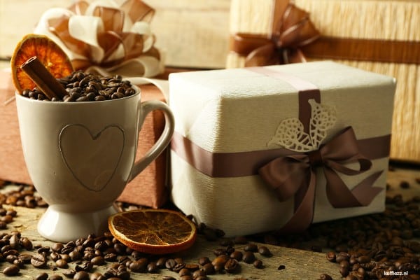 Koffiemok gevuld met koffiebonen naast luxe ingepakte cadeauverpakkingen, omringd door koffiebonen, kaneel en gedroogde sinaasappelschijfjes