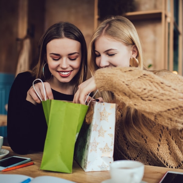 Twee vrouwen kijken enthousiast in cadeautasjes terwijl ze samen cadeaus uitpakken aan tafel