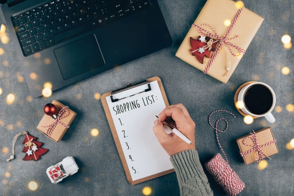 Bovenaanzicht van een bureau met een boodschappenlijstje, ingepakte cadeaus, een laptop en een kop koffie. Een hand schrijft een lijst voor feestelijke aankopen en cadeauvoorbereidingen
