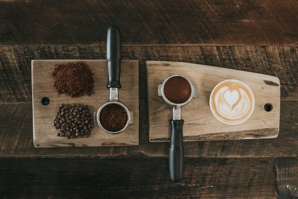 Bovenaanzicht van het koffiezetproces met koffiebonen, gemalen koffie en portafilters naast een cappuccino met latte art op houten serveerplanken