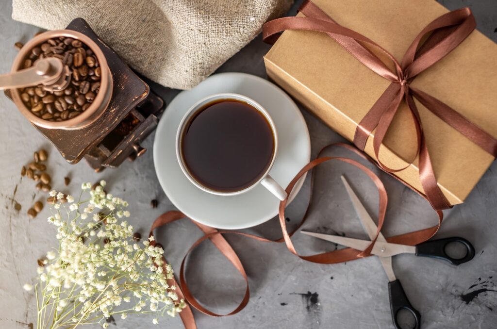 Bovenaanzicht van een kop zwarte koffie naast een ingepakt cadeau, een handmatige koffiemolen en linten