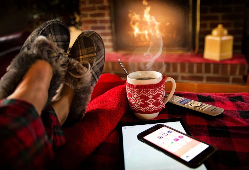 Persoon ontspant bij een knisperende open haard met een warme kop koffie, pantoffels en een deken. Op de voorgrond liggen een smartphone, tablet en afstandsbediening